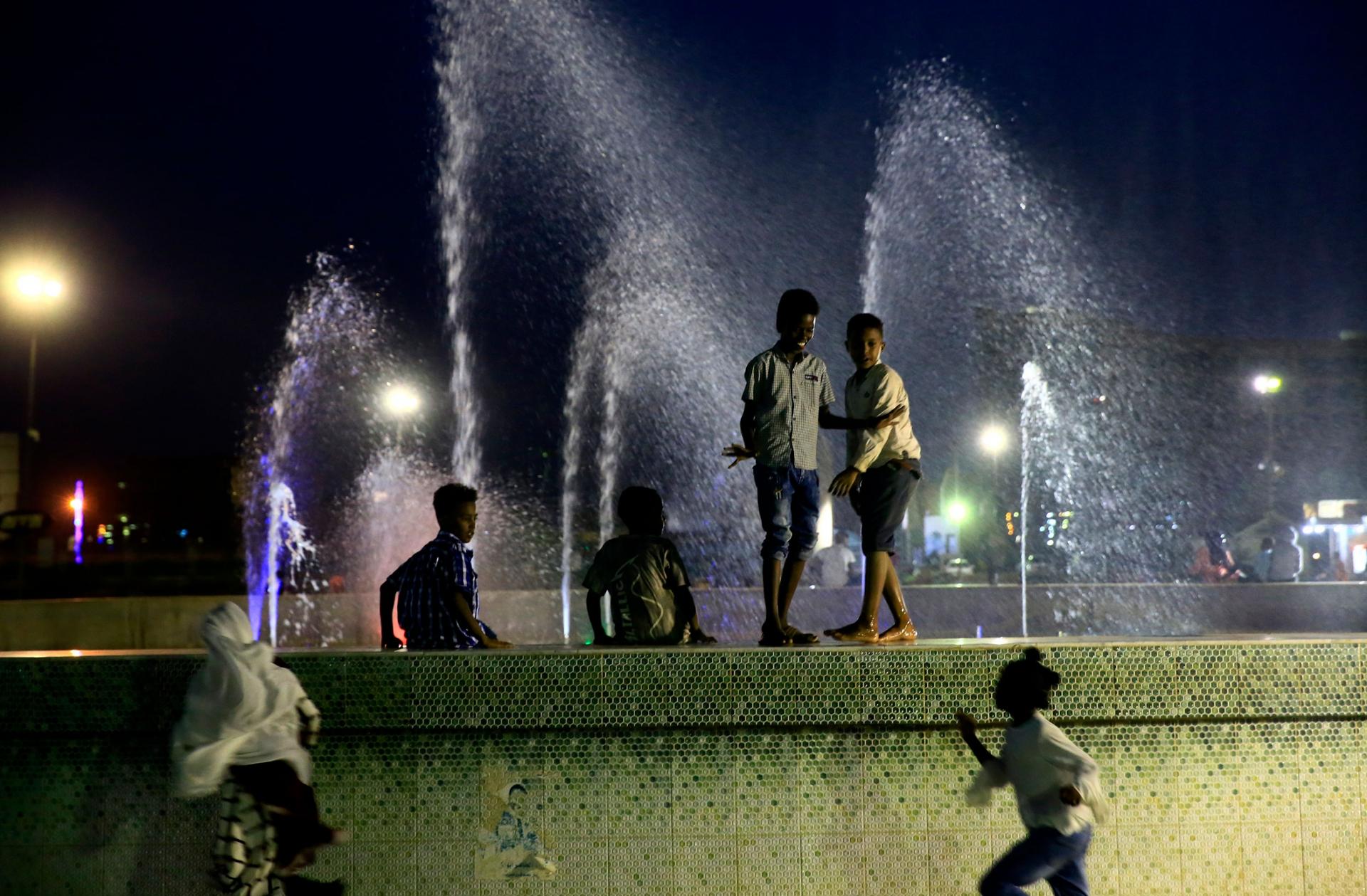 Several children are shown standing on the tiled edge of a fountain with water spraying up in the background.