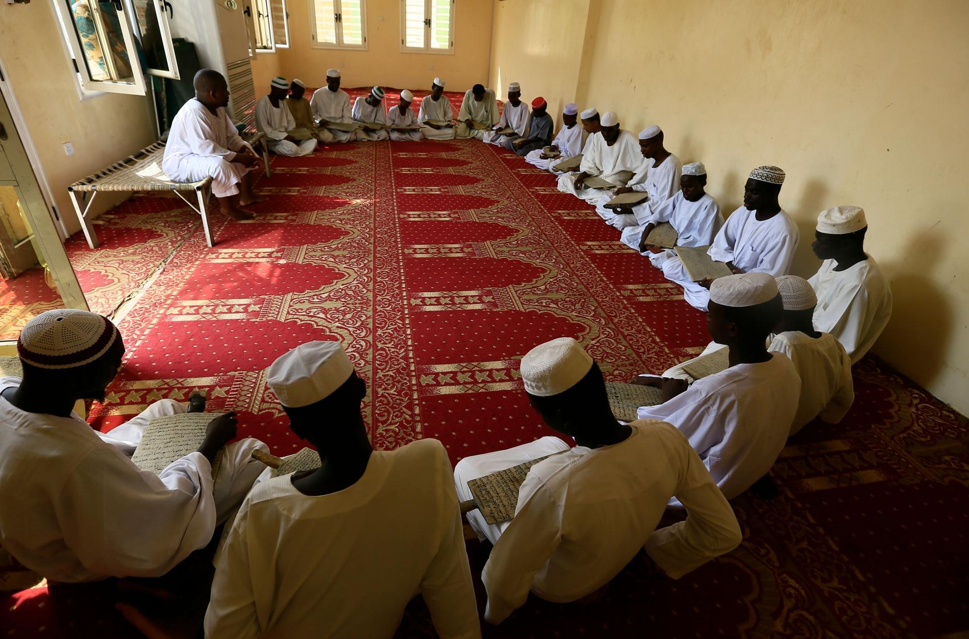 A large group of young men are shown sitting in a half circle on the ground and holding wooden boards with Islamic text.