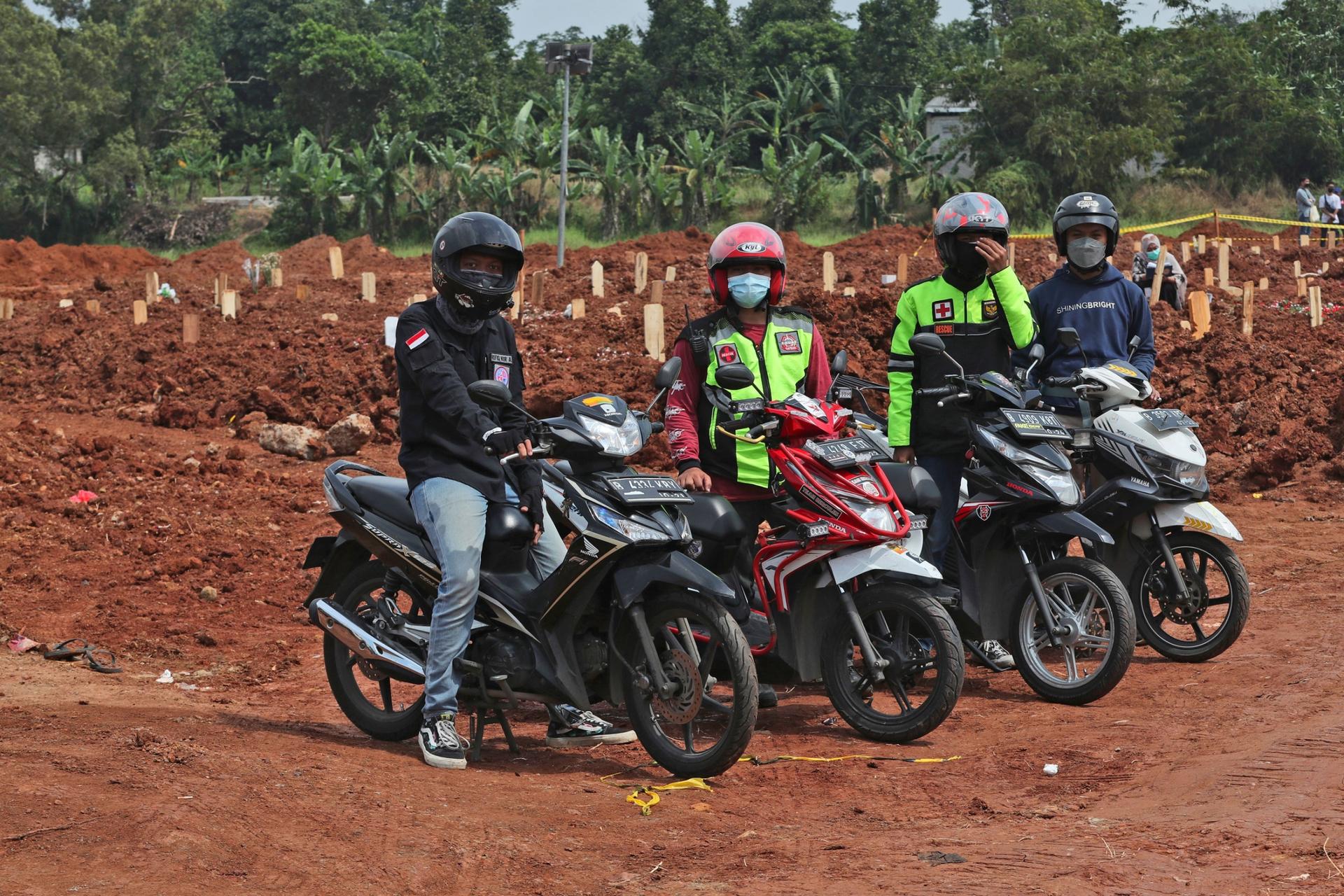 Four people are shown sitting or standing next to motorbikes with a cemetary in the background.
