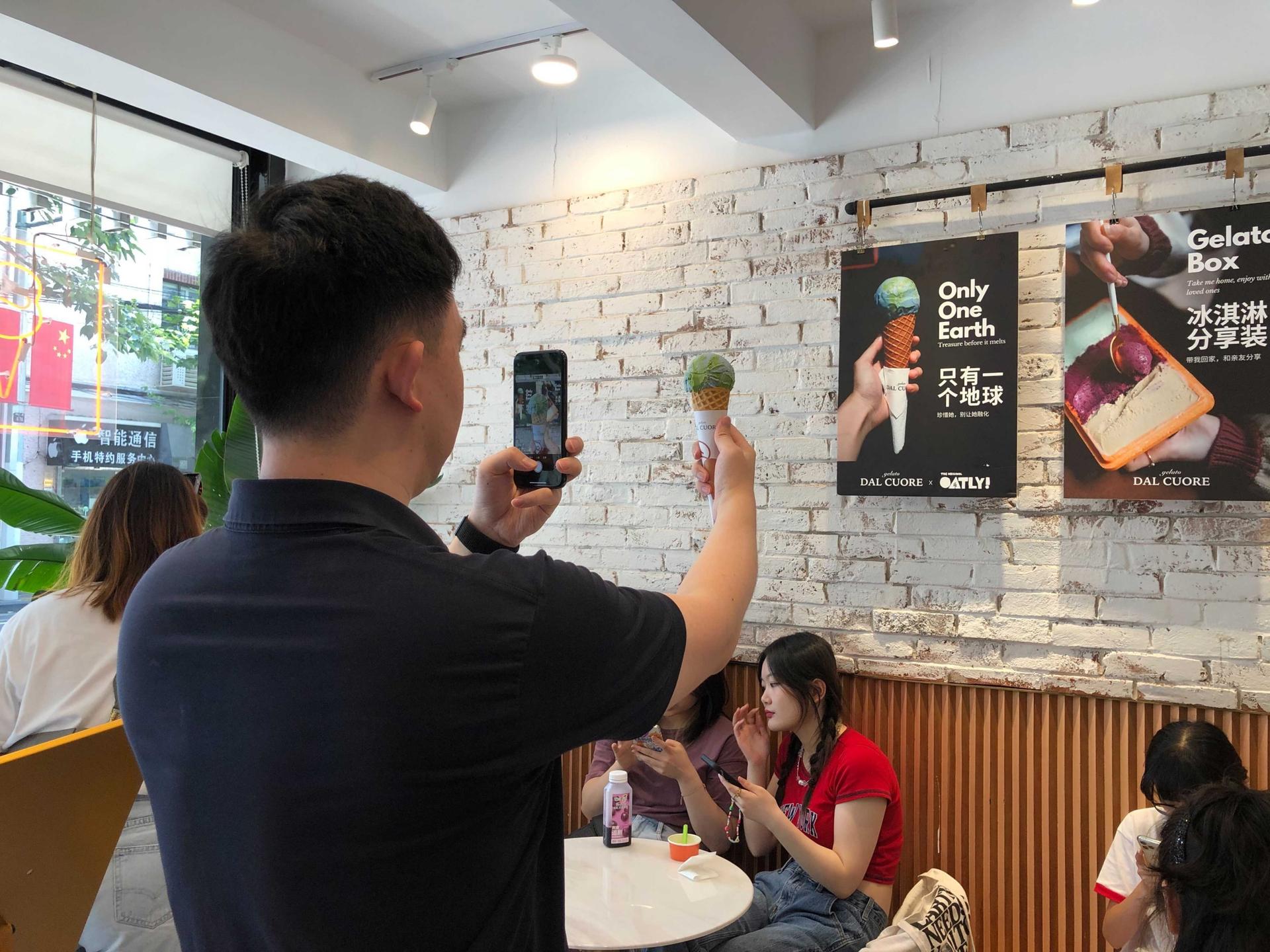 A man holds up a cone with blue and green ice cream as he takes a photo of it against a poster