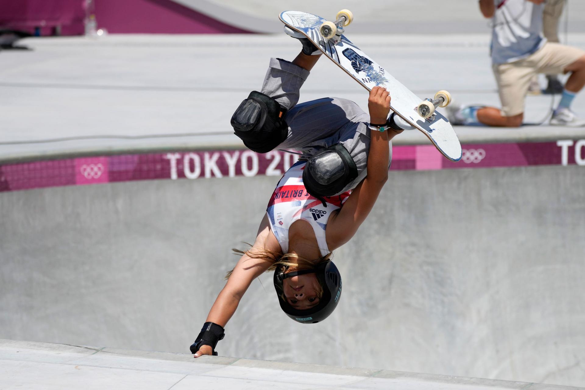 Sky Brown of Britain is shown upside down with her skateboard held against her feet and her right arm holding on to the edge of the park bowl.
