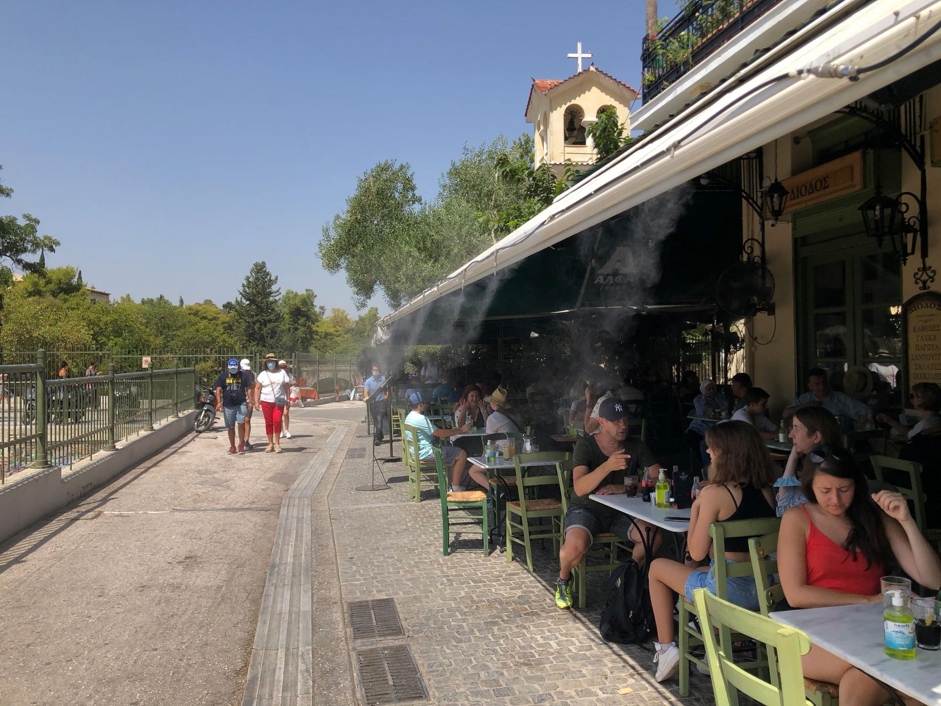 Patrons of a open-air restaurant are shown with several spigots at the roof line spraying mist.