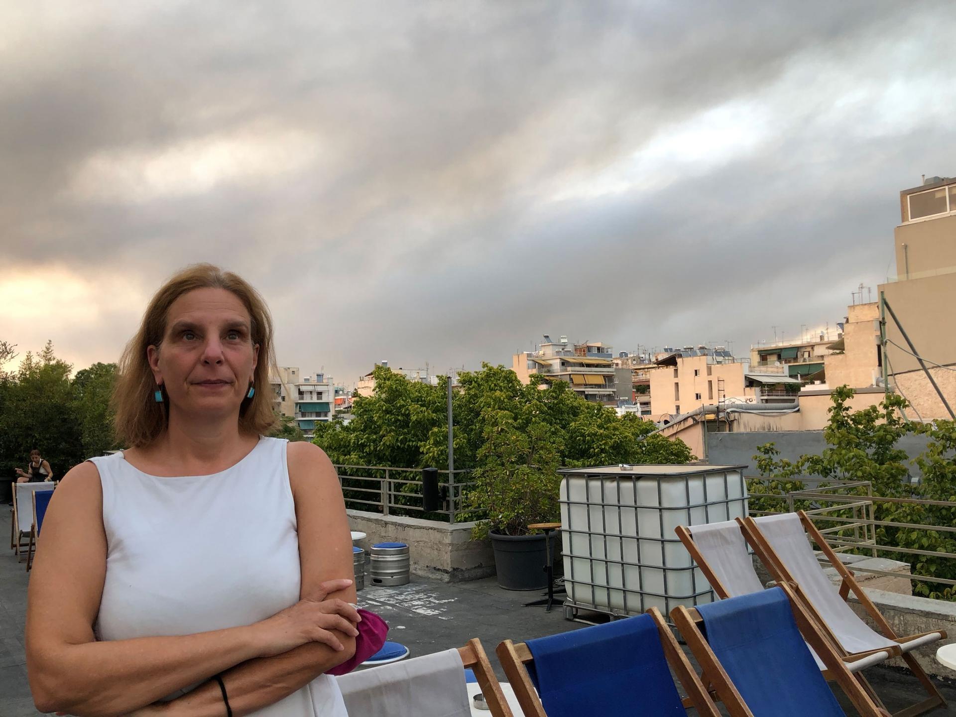 Eleni Myrivili is shown wearing a white sleeveless shirt while standing on a rooftop with the sky behind her filled with smoke clouds.