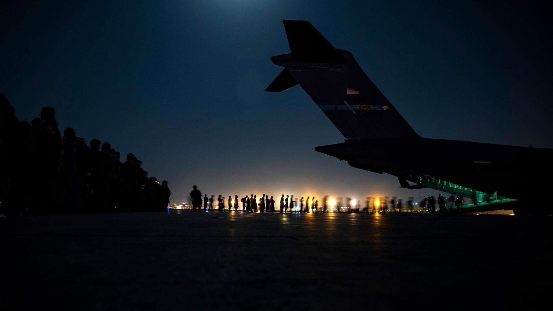A line of people are shown in the distance in low light with the tail of a large aircraft as the sun sets.