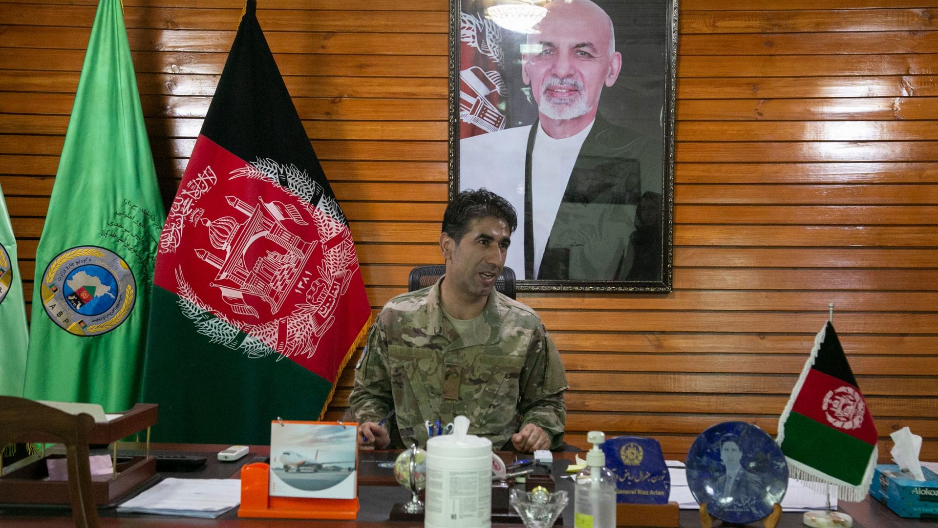 Maj. Gen. Mohammad Reyaz Arian is shown sitting at a large wooden desk and wearing military fatigues with the Afghanistan flag and a portrait photo of former President Ashraf Ghani on the wall.