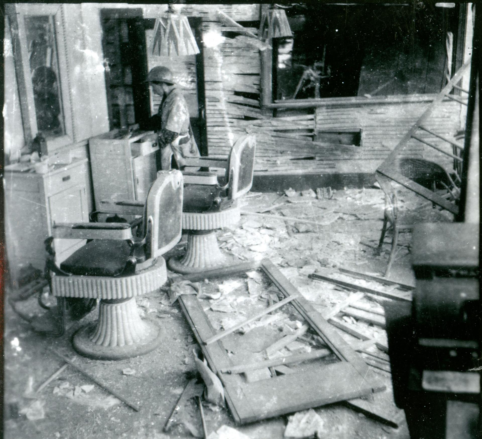 Two barbershop chairs are shown with broken glass on the floor and a woman looking through a drawer.