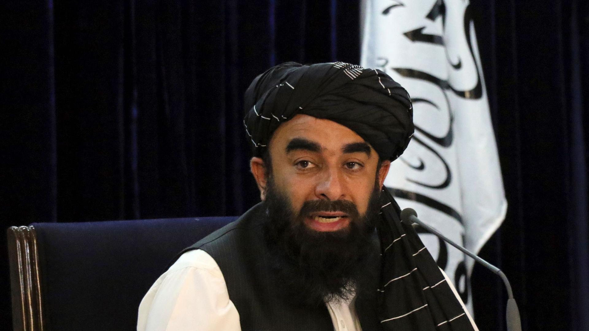 Taliban spokesman Zabihullah Mujahid is shown sitting while wearing a dark head wrap and black vest with the Taliban flag behind him.