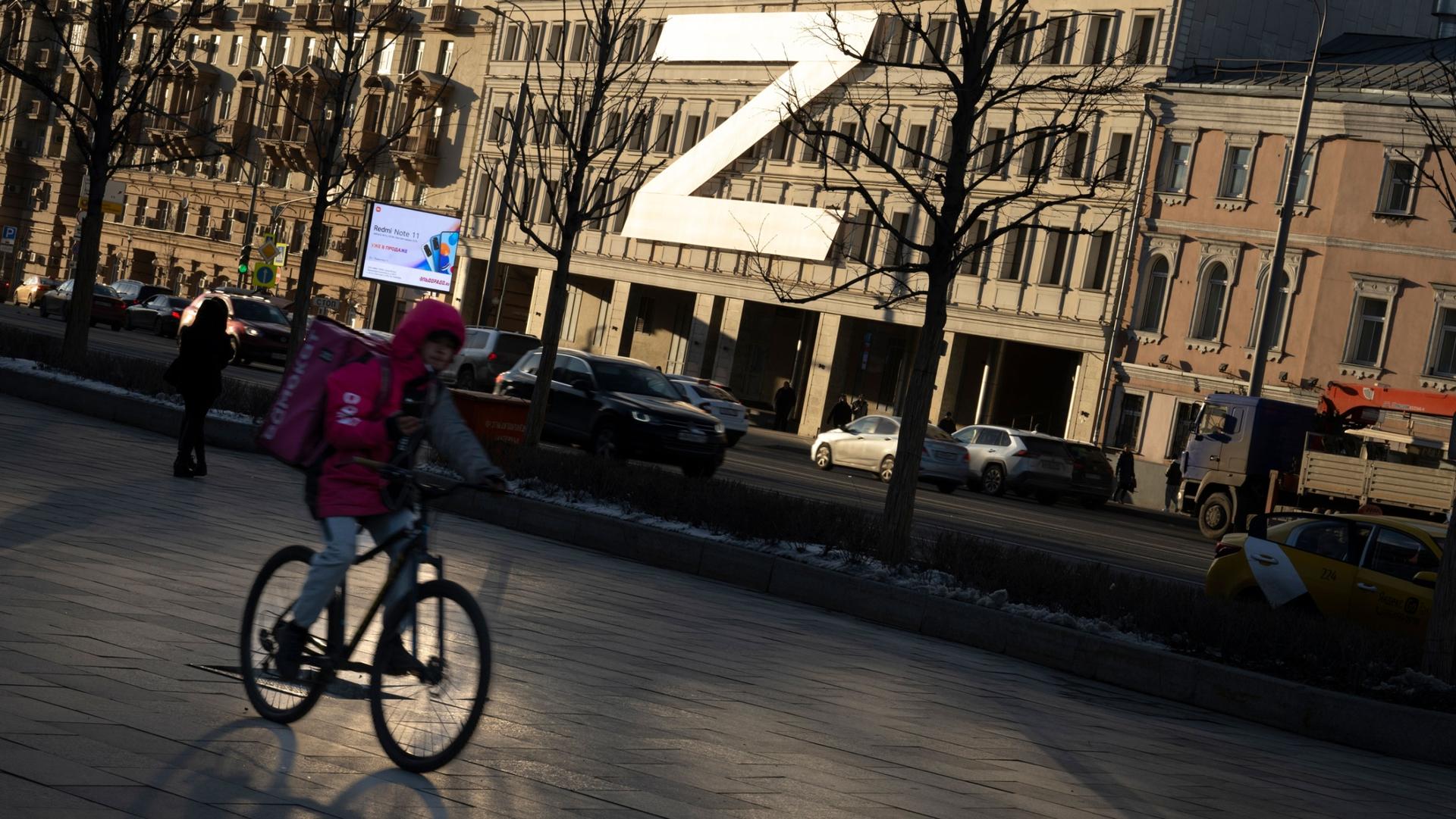 A food delivery courier rides a bicycle along a street with a huge letter Z, which has become a symbol of the Russian military, and a hashtag reading "We don't abandon our own" on a building during sunset in Moscow, Russia, Wednesday, March 30, 2022.