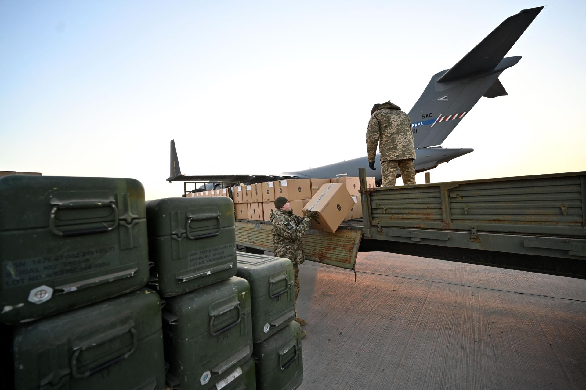 Two soldiers lifting crates onto a military truck with a cargo airplane in the background
