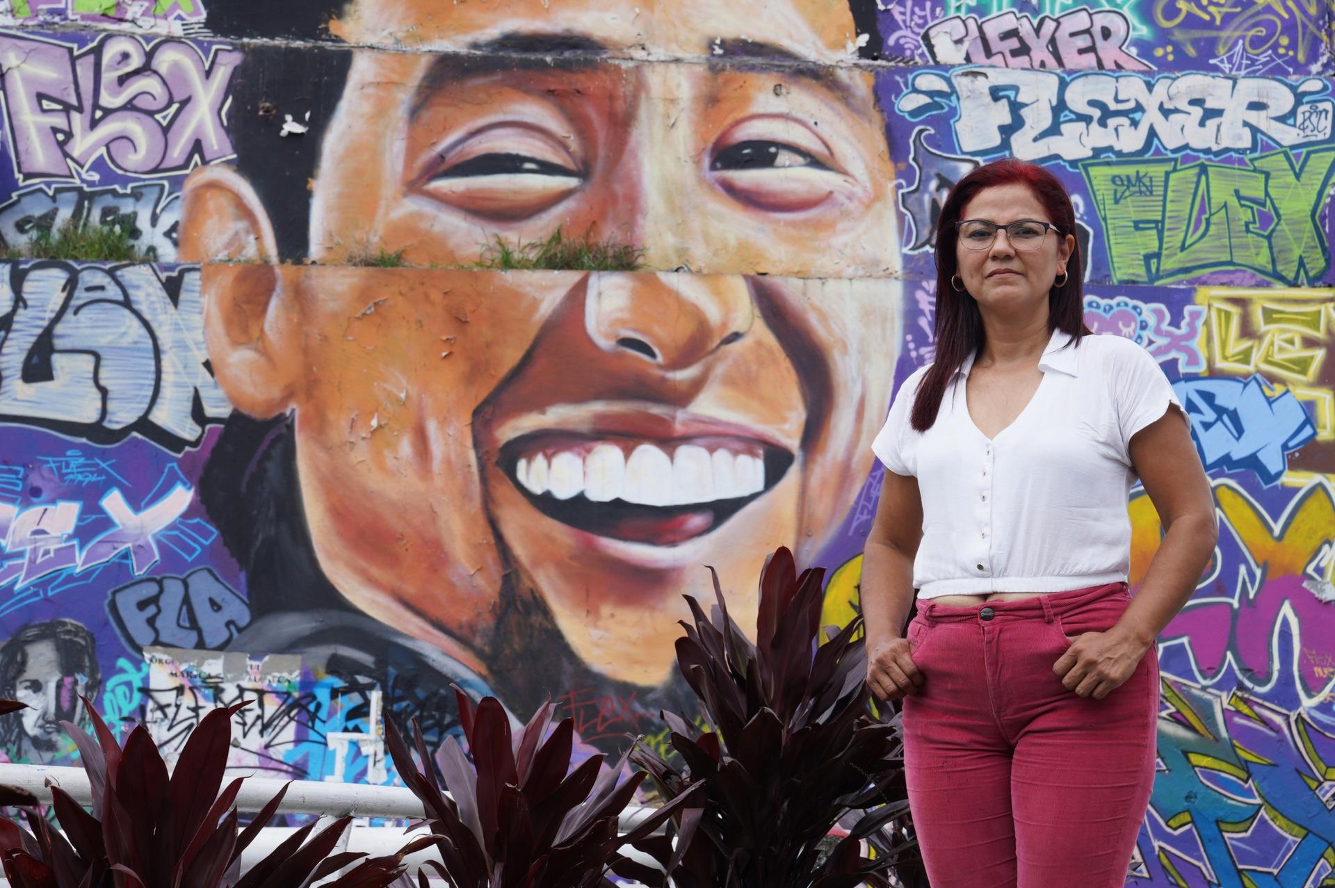 Laura Guerrero, poses for a photo next to a mural in Cali, that depicts her son, Nicolas Guerero