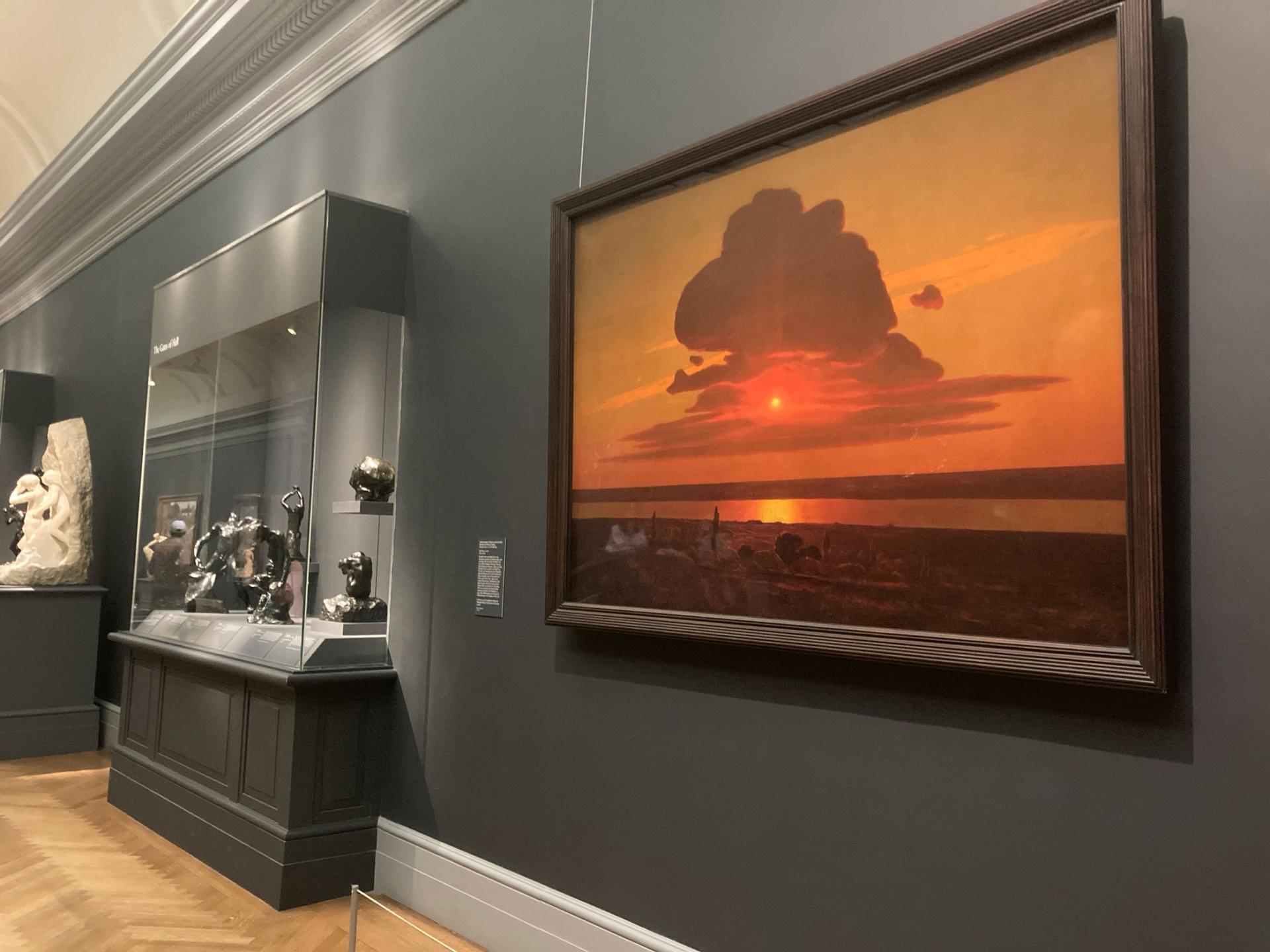 A painting of a red and orange sunset on display against a gray wall near a glass case of sculptures.