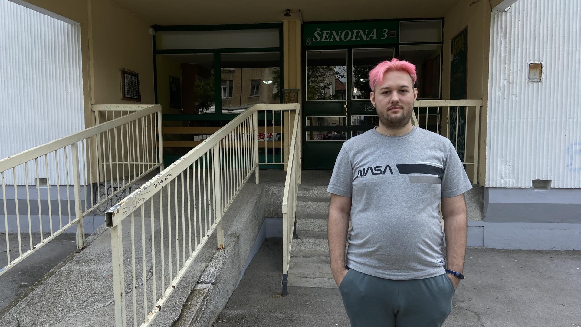 a pink-haired man stands outside the entrance to an apartment building
