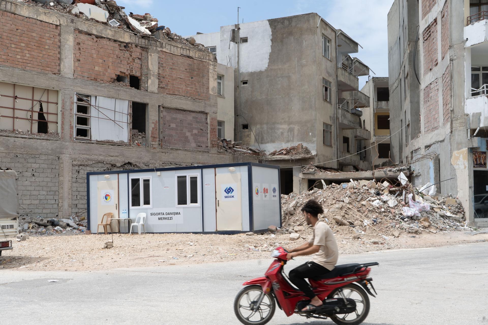 A motorcyclist rides past a demolished building in the beach town of Samandağ. A converted shipping container serves as an insurance office.
