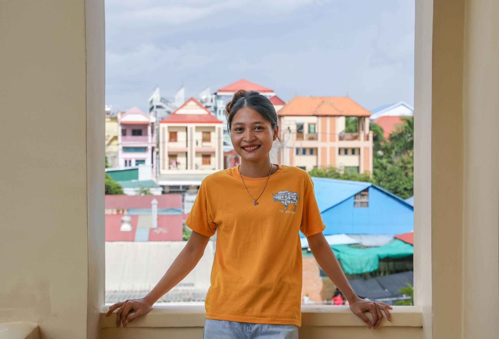 Portrait of a woman in a yellow shirt posing on a balcony. There is a cityscape behind her.