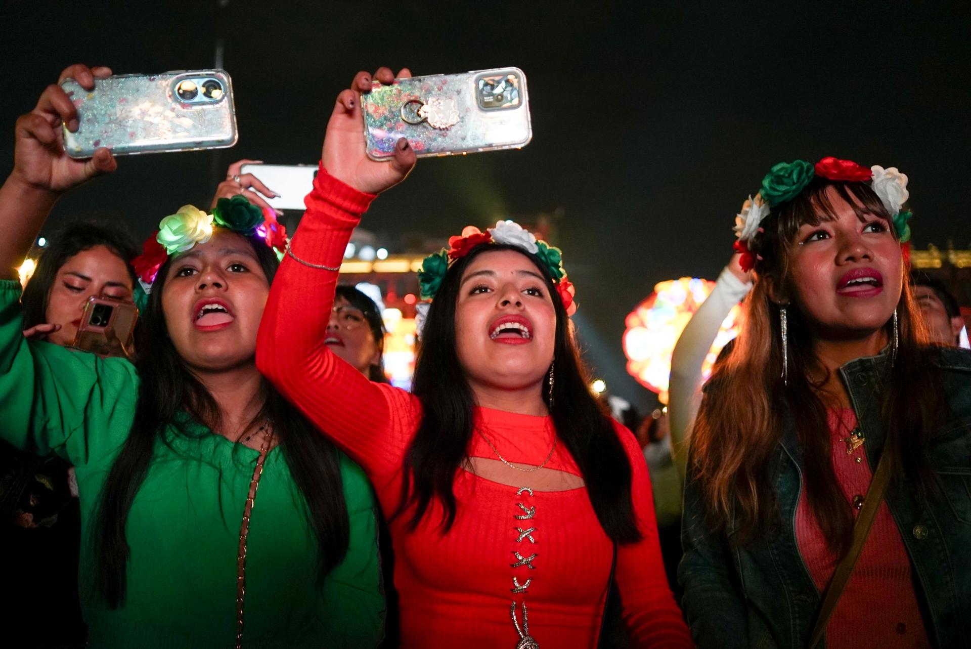 Three young women in red and green celebrate with their phones held up to capture the performance. 