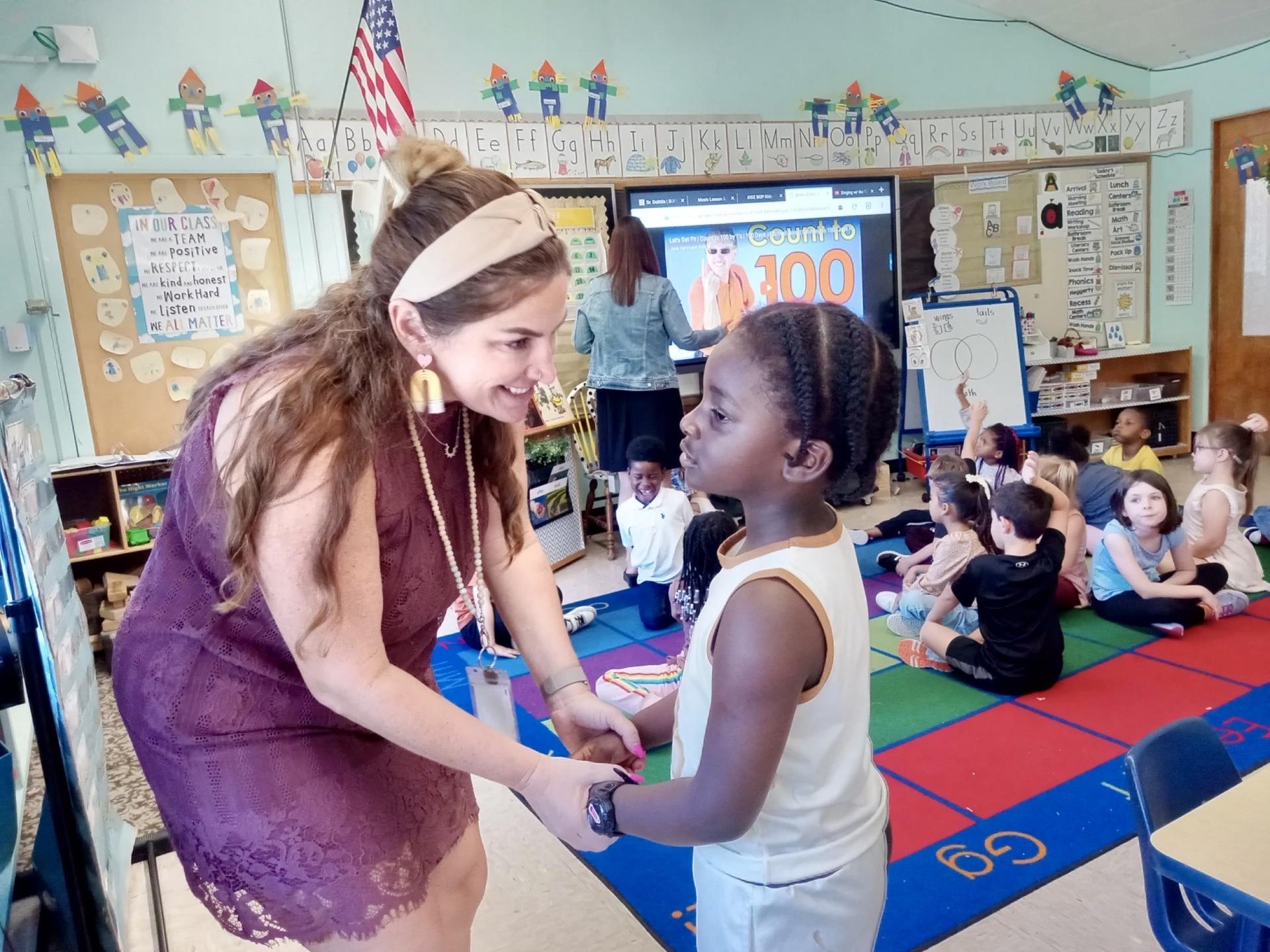 A woman wearing a headband and maroon dress talks to a young child in a busy classroom