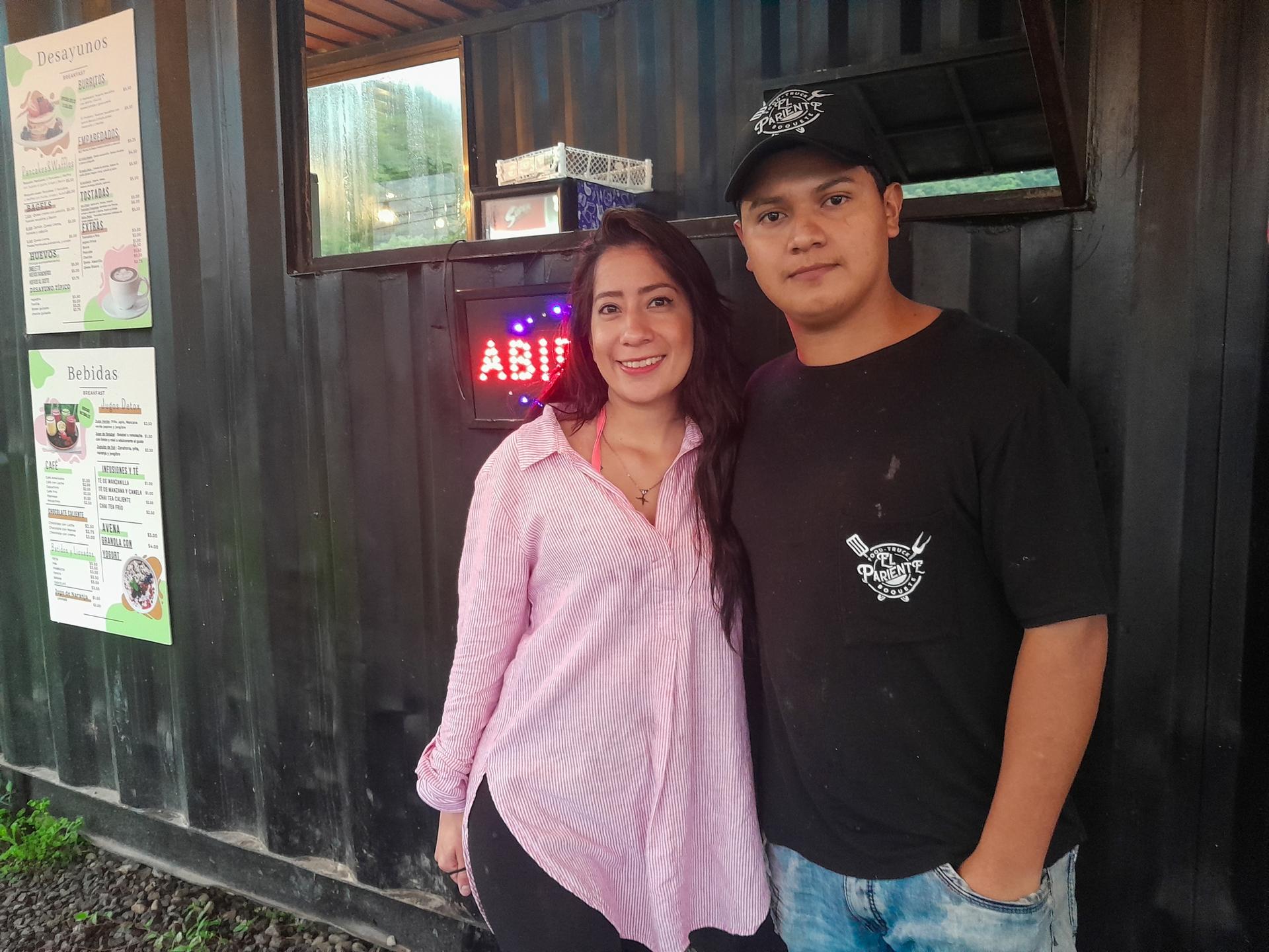 Man and woman stand beside each other posing for photo. The woman wears a pink shirt and the man is wearing a black baseball cap, black shirt and light-colored jeans