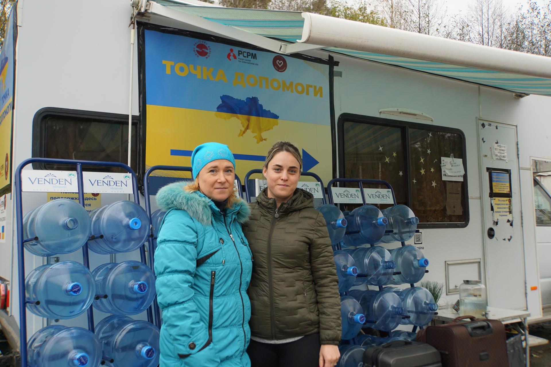 Two women wearing winter coats in front of water jugs