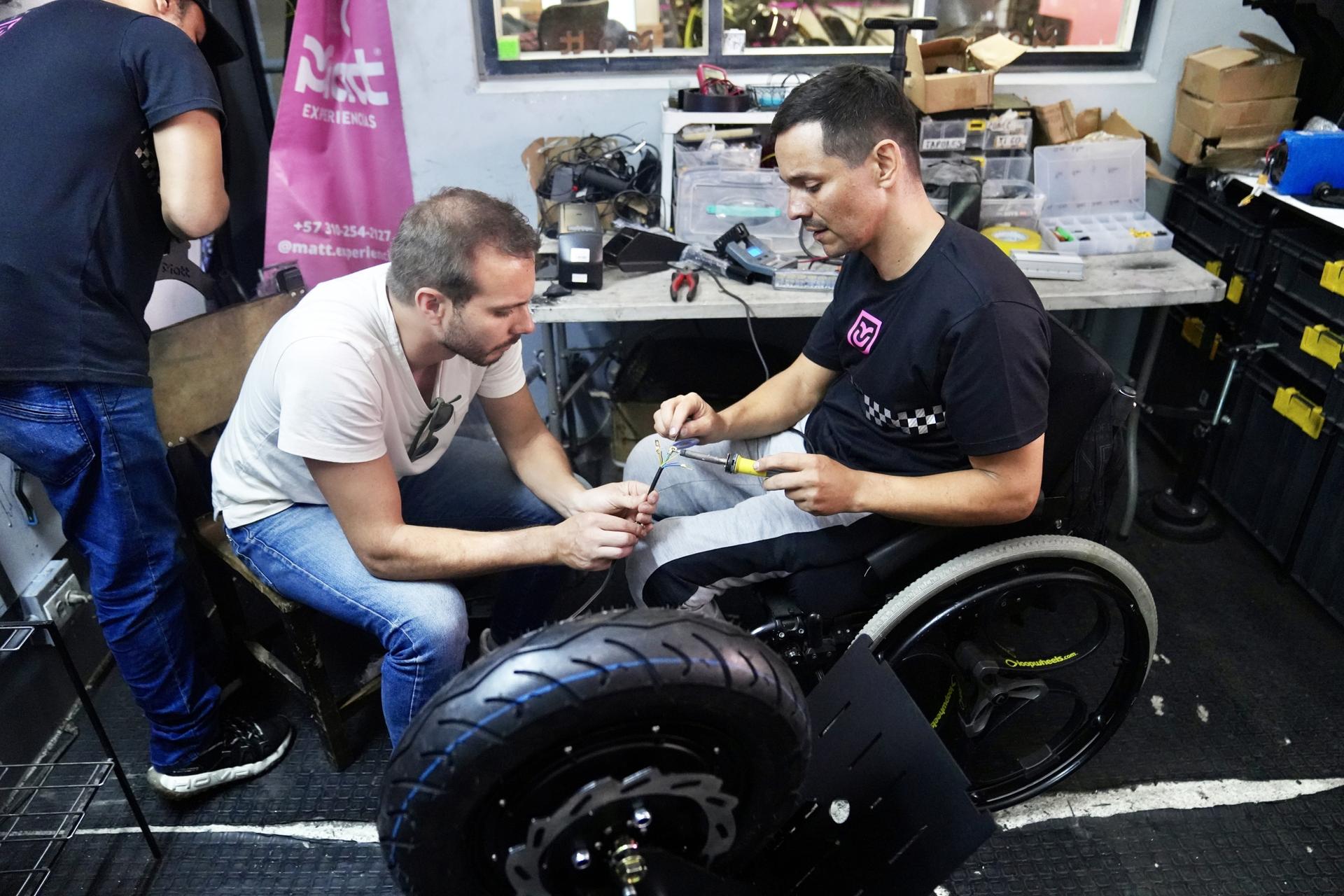 A man in a white shirt and jeans fixing an electric wheelchair that a man in a black shirt is sitting in.