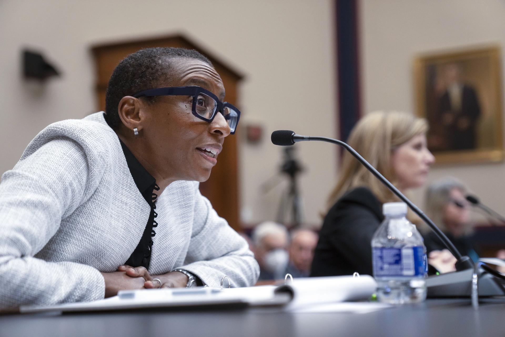 A Black woman wearing glasses and a gray sweater speaking into a microphone in a courtroom