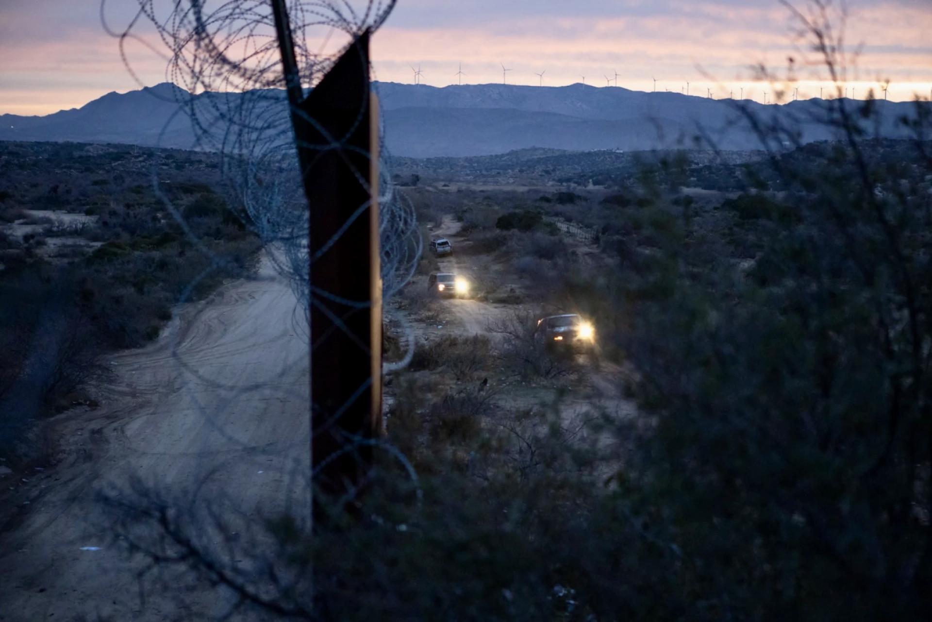 Two trucks filled with migrants approach a gap in the border wall at dawn.