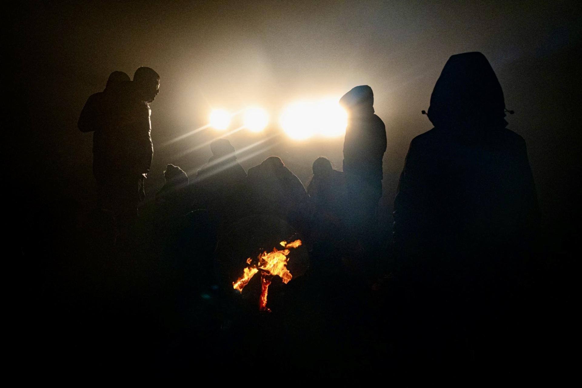 Migrants huddle by a fire as CBP trucks shine their lights on them 