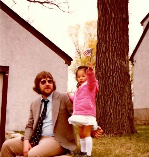 A white man and a small Korean girl in front of a house and tree. The girl waves an American flag. 