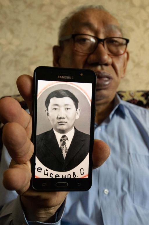 A man wearing glasses holds up a photo of his younger self on his mobile phone. 