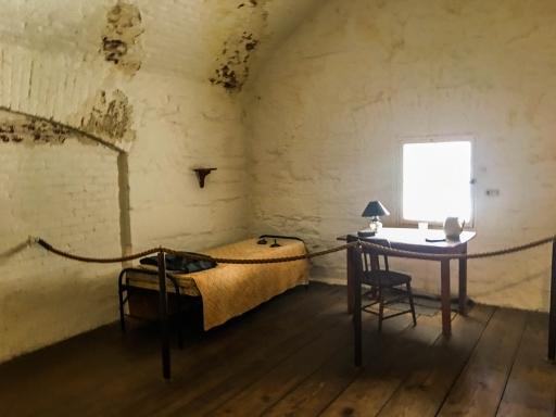 A bed and a desk near a window in a cell. 