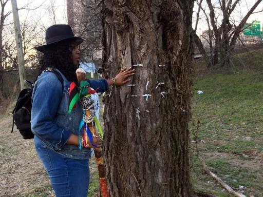 A black woman in a hat stands next to a tree with tags hanging from it. 