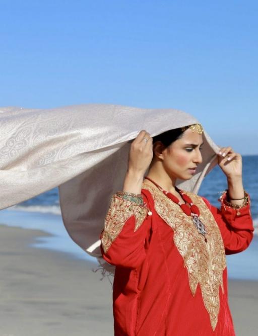 A photograph of a woman in a red pheran holding a scarf that billows out behind her