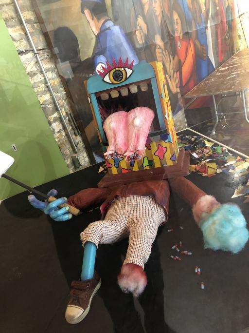 A puppet prop lays on a floor. It has a single eye, and a large pink tongue coming out of a square shaped head