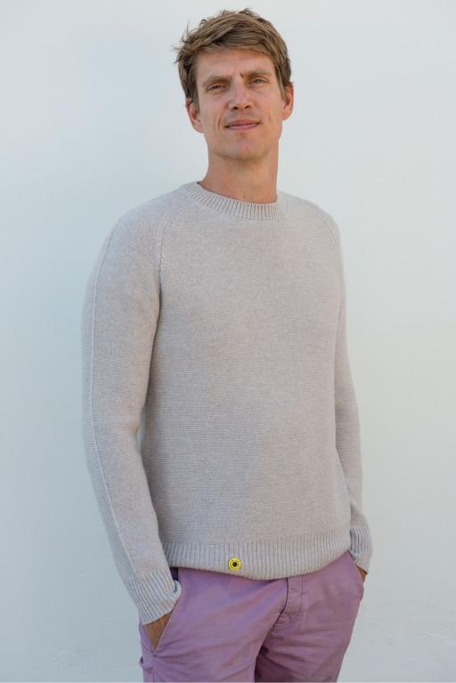 A white man in an off-white sweater and pink pants poses