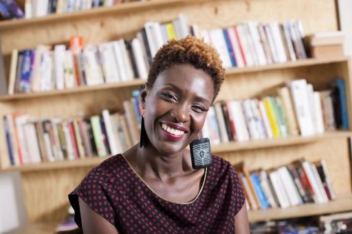 A Black woman with short hair wears earrings and a black and red blouse and smiles warmly in front of a large bookshelf. 