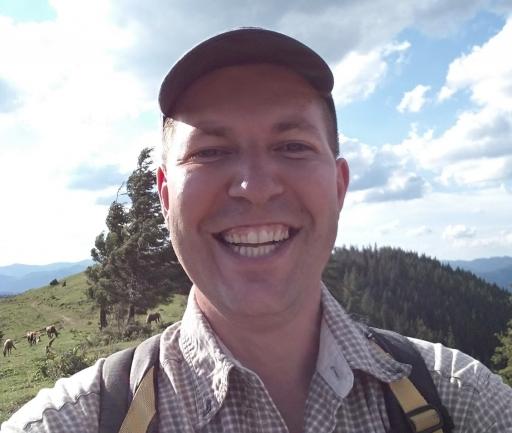 A selfie of Andriy Gnytka, a white man wearing a hat and smiling in the sunshine with his backpack on his back and trees behind him