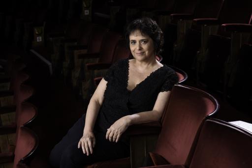 A white woman with dark black hair wearing all black clothing poses seated in a chair at a theater, smiling to the camera with her arms on her lap. 