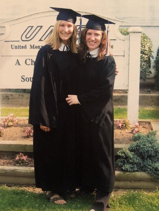 Two young women wear graduation robes and caps and stand close for a photo. 