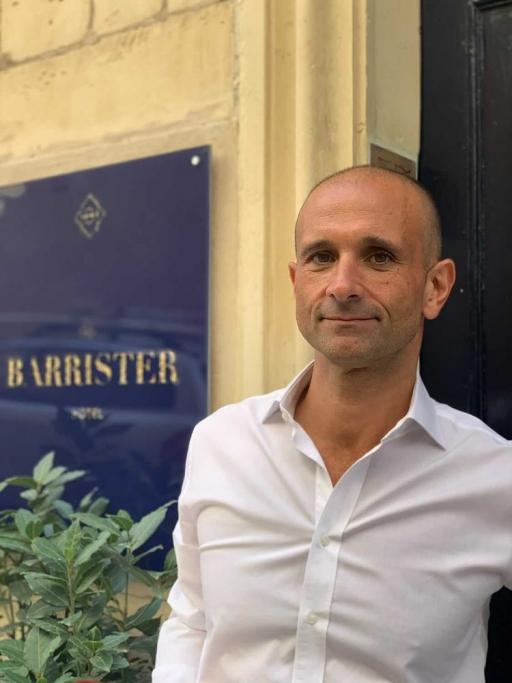 Mark Gauci wears a white button-down shirt near a blue sign that reads "The Barrister," his hotel. 