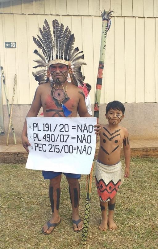An Indigenous man wearing a feathered crown stands near a small boy and holds a sign. 