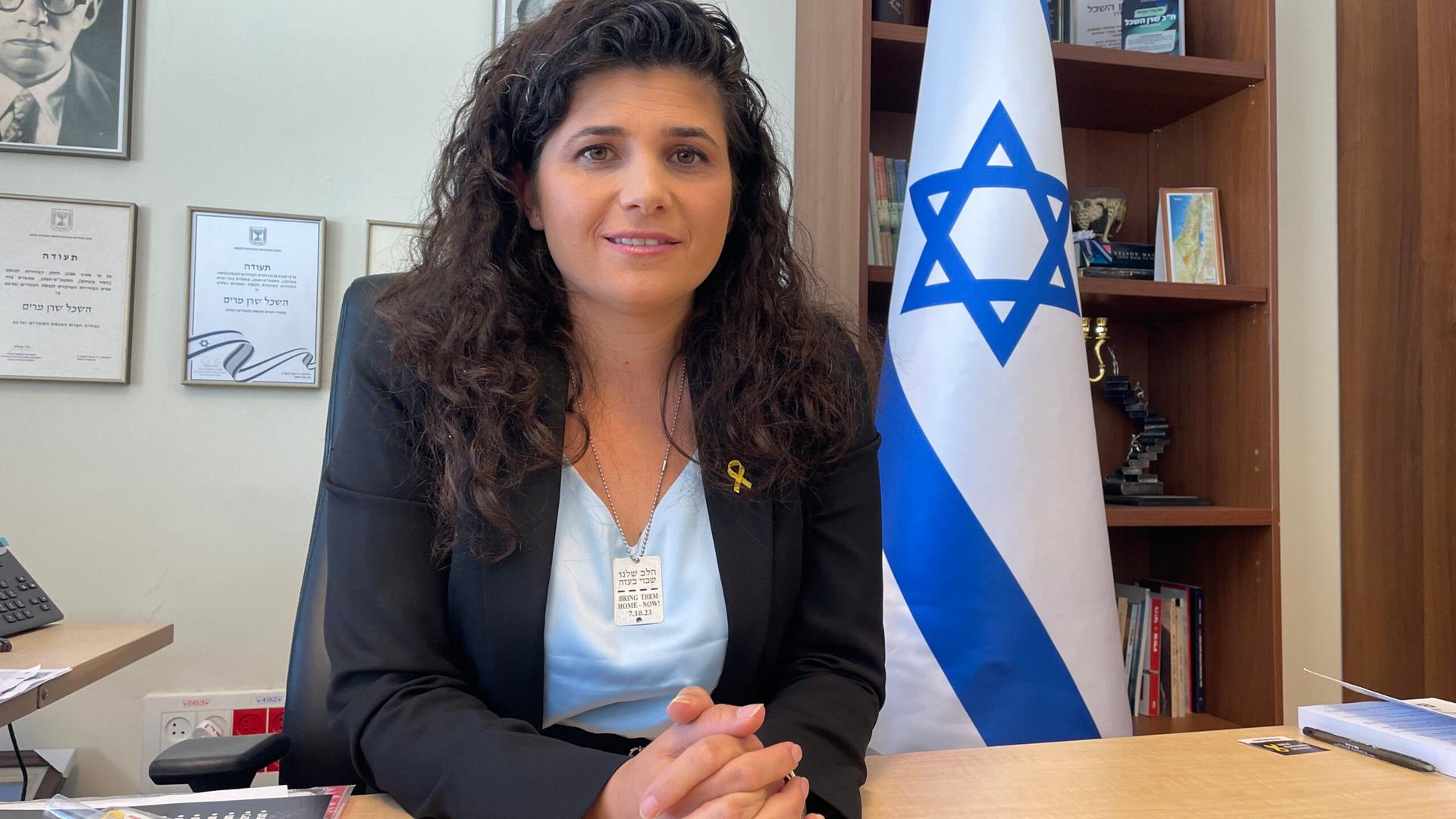A woman wearing a light blue blouse with a black blazer sitting down next to an Israeli flag
