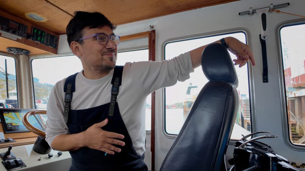 A man in a boat cabin wearing overalls and glasses, smiling while resting his arm on the back of a driver's seat.