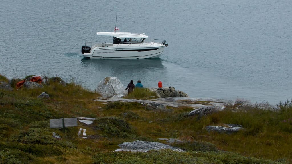A motorboat labeled 'Askeladden P76 Weekend' on a body of water near a rocky shore with two people wearing coats sitting on the rocks, surrounded by grass and small shrubs.