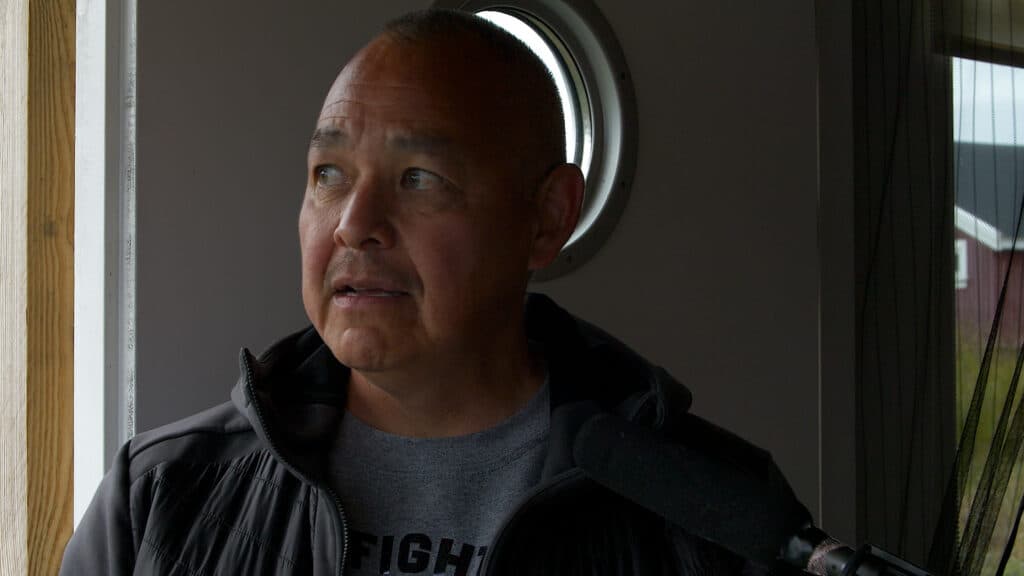 A person standing indoors, looking slightly to the side, with a circular window behind them and a microphone visible in the foreground.