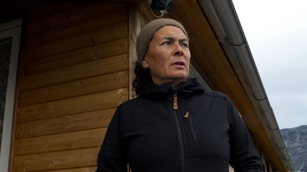 A person wearing a black jacket and a brown headband stands in front of a wooden cabin, looking to the side.