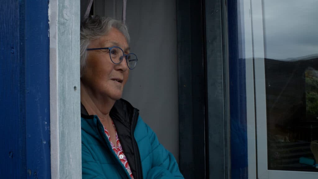 Elderly person with grey hair and round glasses looks out from a window, wearing a blue jacket and standing in a doorway of a blue-painted structure.