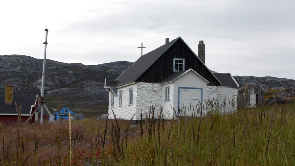 A small white chapel with blue trim and a black roof, featuring a cross on top, situated in a grassy field with rocky hills in the background.