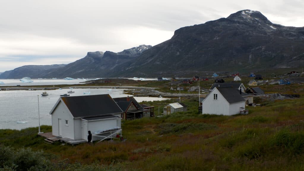 Coastal village with small colorful houses, a white wooden building in the foreground, and icebergs floating in the sea, set against a backdrop of rugged mountains under a cloudy sky.