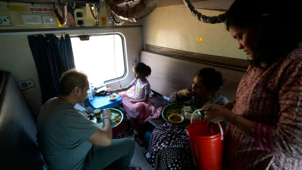 A family dining inside a train compartment, with a man, a woman, and a child seated around a small table enjoying a meal. Another woman is standing, holding an orange container. The compartment has bunk beds and a window with curtains partially drawn.