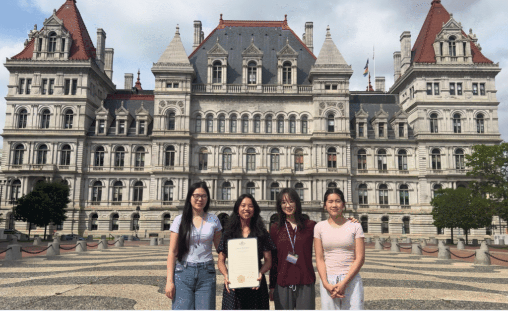 Four people standing in front of a historic building with ornate architecture, featuring pointed roofs and a symmetrical facade. One person is holding a certificate. The group is positioned on a patterned courtyard with greenery in the background.