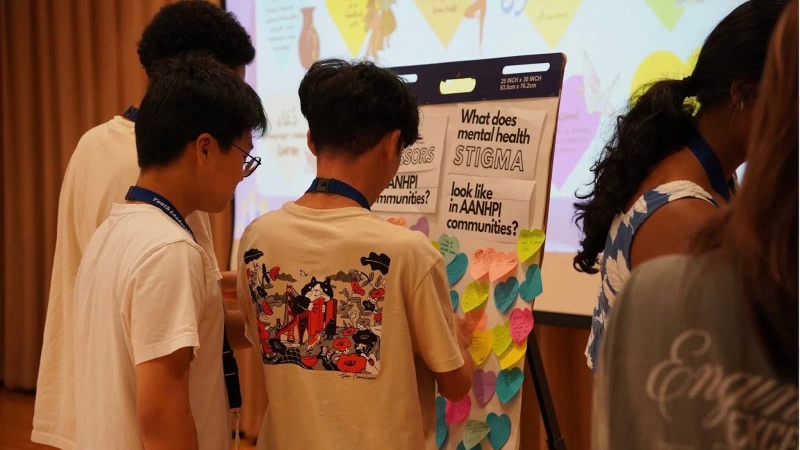 A group of young people standing around a flip chart discussing mental health stigma in AANHPI communities. The chart is covered with colorful heart-shaped notes with handwritten messages. They are in an indoor setting, possibly a workshop or seminar.